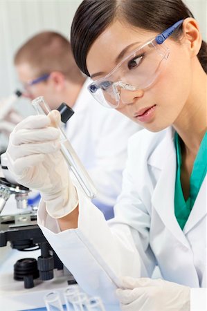 A Chinese Asian female medical or scientific researcher or doctor using looking at a test tube of clear liquid in a laboratory with her colleague out of focus behind her. Stock Photo - Budget Royalty-Free & Subscription, Code: 400-05909911