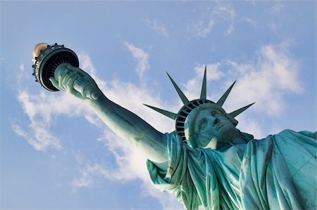 estatua de la libertad - Unique perspective image of the upper torso, head, arm and torch of the Statue of Liberty.  The image is shot against a slightly cloudy blue sky. Foto de stock - Super Valor sin royalties y Suscripción, Código: 400-06366088