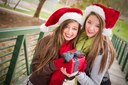 Two Attractive Festive Smiling Mixed Race Women Wearing Christmas Santa Hats Holding a Wrapped Gift with Bow Outside. Photographie de stock - Aubaine LD & Abonnement, Code: 400-06465320