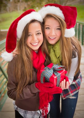 Two Attractive Festive Smiling Mixed Race Women Wearing Christmas Santa Hats Holding a Wrapped Gift with Bow Outside. Photographie de stock - Aubaine LD & Abonnement, Code: 400-06465321
