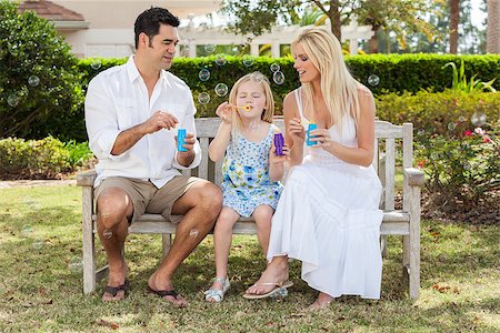 A young family mother & father parents with girl child blowing bubbles having fun together sitting on a bench in a sunny park or garden. Stock Photo - Budget Royalty-Free & Subscription, Code: 400-06562527