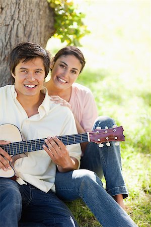Woman smiling while resting her hand on the shoulder of her friend who i holding a guitar as they both sit against a tree Stock Photo - Budget Royalty-Free & Subscription, Code: 400-06635936