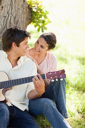 Smiling woman looking eye to eye with her friend who is holding a guitar as they both sit against a tree Stock Photo - Budget Royalty-Free & Subscription, Code: 400-06635937