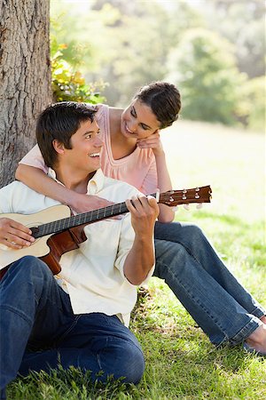 Woman looking at her friend and resting her arm on his chest while he plays a guitar under a tree Foto de stock - Super Valor sin royalties y Suscripción, Código: 400-06635931