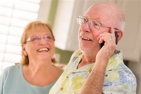 simsearch:400-04522125,k - Happy Senior Adult Husband on Cell Phone with Wife Behind in Kitchen. Stock Photo - Budget Royalty-Free & Subscription, Code: 400-06701036