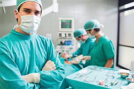 simsearch:400-06735487,k - Surgeon with arms crossed looking at camera with colleagues performing in background Fotografie stock - Microstock e Abbonamento, Codice: 400-06735481