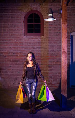 Excited Pretty Mixed Race Young Adult Woman Walking in the Evening Holding Shopping Bags with Brick Wall Background. Stock Photo - Budget Royalty-Free & Subscription, Code: 400-06794558