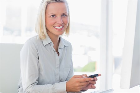 simsearch:400-06883455,k - Blonde businesswoman texting and smiling at camera at her desk Photographie de stock - Aubaine LD & Abonnement, Code: 400-06884364