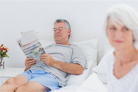 simsearch:400-06888132,k - Mature man reading a newspaper in bed while his wife is sitting on bed Photographie de stock - Aubaine LD & Abonnement, Code: 400-06888135