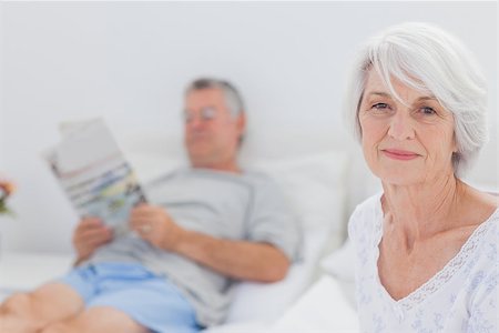 simsearch:400-06888132,k - Smiling mature woman sitting in bed with husband reading a newspaper on the background Photographie de stock - Aubaine LD & Abonnement, Code: 400-06888136