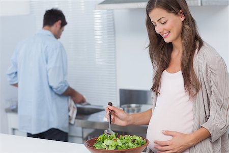 simsearch:6109-08488688,k - Pregnant woman mixing a salad in the kitchen while her husband is washing the dishes behind Stock Photo - Budget Royalty-Free & Subscription, Code: 400-06888618