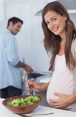 simsearch:6109-08488688,k - Pregnant woman preparing a salad in the kitchen with her husband washing the dishes behind Photographie de stock - Aubaine LD & Abonnement, Code: 400-06888619