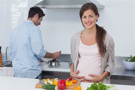 pregnant woman cooking photos - Pregnant woman holding her belly in the kitchen with her husband cooking behind Stock Photo - Budget Royalty-Free & Subscription, Code: 400-06888626