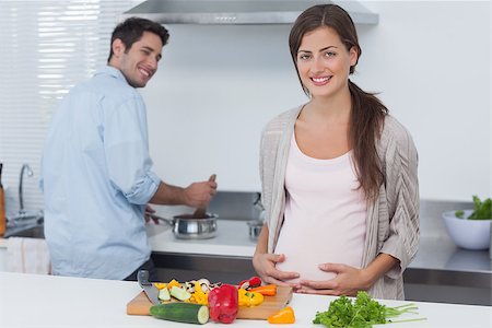 pregnant woman cooking photos - Cheerful pregnant woman holding her belly in the kitchen with her husband cooking behind Stock Photo - Budget Royalty-Free & Subscription, Code: 400-06888627