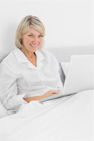 simsearch:400-06891126,k - Blonde woman sitting in bed with laptop smiling at camera at home in bedroom Stock Photo - Budget Royalty-Free & Subscription, Code: 400-06891101