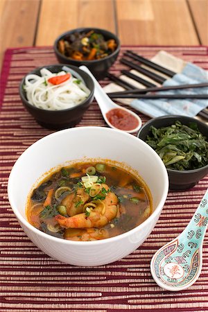 Asian soup with shrimps in white ceramic bowl composed with ceramic spoon with spicy red sauce, asian chopsticks and black bowls with rice noodles, kale (green cabbage) and fried vegetables. Composition on a old styled wooden table. Stock Photo - Budget Royalty-Free & Subscription, Code: 400-06912832