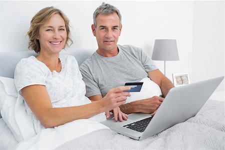 Smiling couple using their laptop to buy online in bed looking at camera at home in bedroom Stock Photo - Budget Royalty-Free & Subscription, Code: 400-06931280