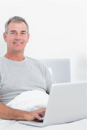 Smiling grey haired man using his laptop in bed looking at camera in bedroom at home Stock Photo - Budget Royalty-Free & Subscription, Code: 400-06931325