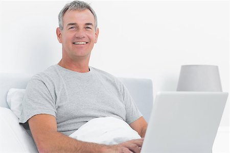 Happy grey haired man using his laptop in bed smiling at camera in bedroom at home Stock Photo - Budget Royalty-Free & Subscription, Code: 400-06931323