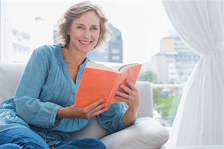simsearch:400-06872352,k - Cheerful blonde woman sitting on her couch holding a book smiling at camera at home in the sitting room Foto de stock - Royalty-Free Super Valor e Assinatura, Número: 400-06931339