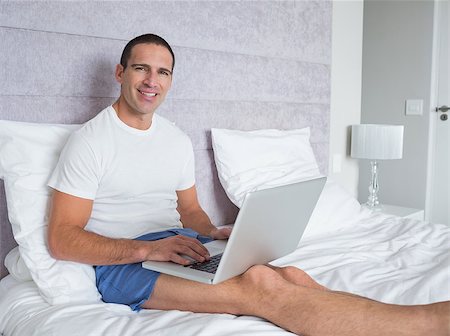 Happy man using laptop on bed smiling at camera at home in the bedroom Stock Photo - Budget Royalty-Free & Subscription, Code: 400-06931580