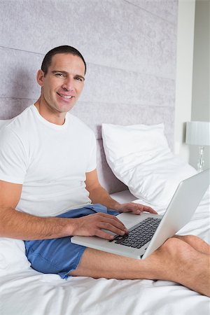 Smiling man using laptop on bed looking at camera at home in the bedroom Stock Photo - Budget Royalty-Free & Subscription, Code: 400-06931581
