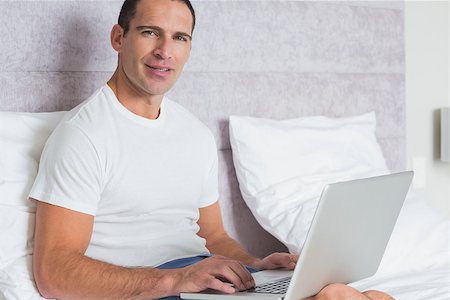 Cheerful man using laptop on bed smiling at camera at home in the bedroom Stock Photo - Budget Royalty-Free & Subscription, Code: 400-06931583