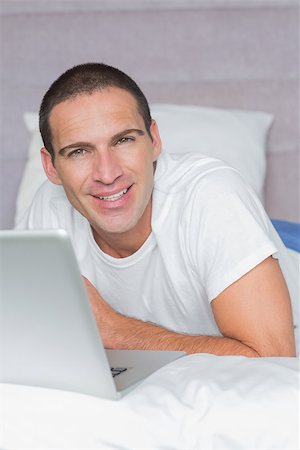 simsearch:6109-08489191,k - Cheerful man lying on bed using his laptop looking at camera at home in the bedroom Fotografie stock - Microstock e Abbonamento, Codice: 400-06931598