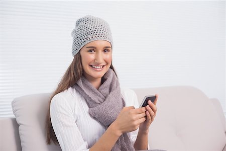 Smiling cute brunette with winter hat on using her smartphone sitting on cosy sofa Stock Photo - Budget Royalty-Free & Subscription, Code: 400-06956156