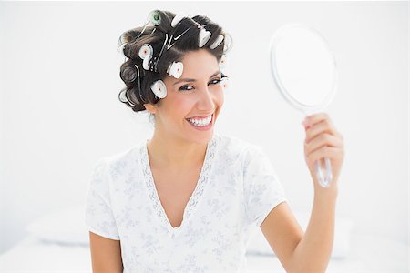 Happy brunette in hair rollers holding hand mirror smiling at camera at home in bedroom Stock Photo - Budget Royalty-Free & Subscription, Code: 400-06958199