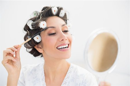 Smiling brunette in hair rollers looking in hand mirror and brushing her eyebrows at home in bedroom Photographie de stock - Aubaine LD & Abonnement, Code: 400-06958214