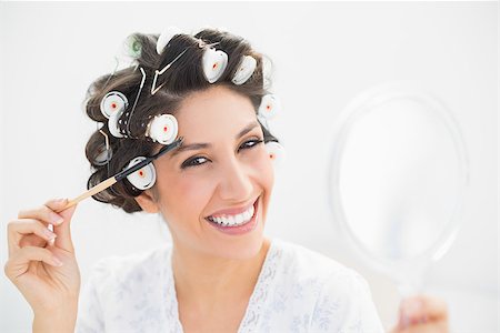 Pretty brunette in hair rollers holding hand mirror and brushing her eyebrows in bedroom at home Stock Photo - Budget Royalty-Free & Subscription, Code: 400-06958215