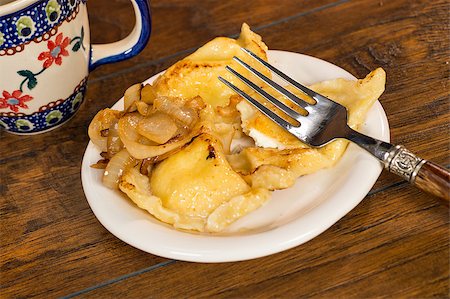 A plate of handmade pierogi with a side of caramelized onions.  There is a decorative silver and wood fork on the plate, as well as a colorful mug in the background.  Everything is shot on a wood table. Photographie de stock - Aubaine LD & Abonnement, Code: 400-07043696