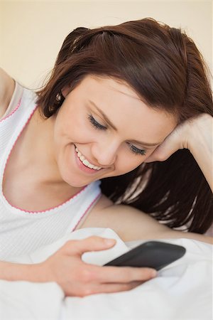simsearch:400-06932687,k - Cheerful brunette looking and using her phone lying on a bed in a bedroom Stock Photo - Budget Royalty-Free & Subscription, Code: 400-07059103