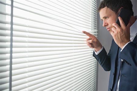 Unsmiling handsome businessman looking through roller blind phoning in dark room Stock Photo - Budget Royalty-Free & Subscription, Code: 400-07139147