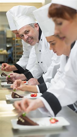 simsearch:400-07140008,k - Team of chefs in a row garnishing dessert plates one smiling at camera in busy kitchen Photographie de stock - Aubaine LD & Abonnement, Code: 400-07139981