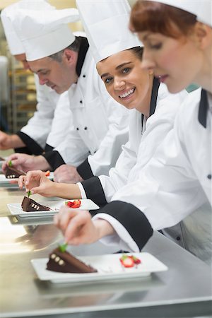 simsearch:400-07140008,k - Team of chefs in a row garnishing dessert plates one girl smiling at camera in busy kitchen Photographie de stock - Aubaine LD & Abonnement, Code: 400-07139982