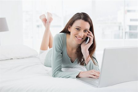 Portrait of a smiling casual young woman using cellphone and laptop in bed at home Stock Photo - Budget Royalty-Free & Subscription, Code: 400-07178628