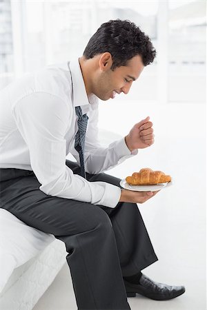 sophisticated home smile - Side view of a smiling young businessman sitting on bed with a plate of croissants at home Stock Photo - Budget Royalty-Free & Subscription, Code: 400-07228472