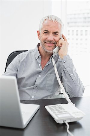 simsearch:400-07230809,k - Portrait of a smiling businessman using telephone in front of laptop at office desk Foto de stock - Super Valor sin royalties y Suscripción, Código: 400-07230829