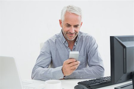 simsearch:400-07230809,k - Smiling mature businessman with cellphone, laptop and computer at desk against white background Foto de stock - Super Valor sin royalties y Suscripción, Código: 400-07230835