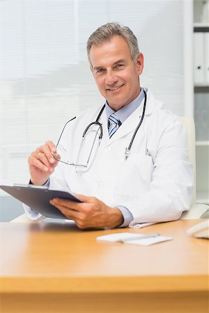 simsearch:614-06898452,k - Smiling doctor sitting at his desk with clipboard his office at the hospital Stock Photo - Budget Royalty-Free & Subscription, Code: 400-07275587