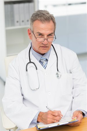 simsearch:614-06898452,k - Serious doctor sitting at his desk writing on clipboard in his office at the hospital Stock Photo - Budget Royalty-Free & Subscription, Code: 400-07275588