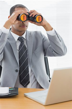 simsearch:400-07266908,k - Serious young businessman looking through binoculars in front of laptop at office desk Photographie de stock - Aubaine LD & Abonnement, Code: 400-07272729