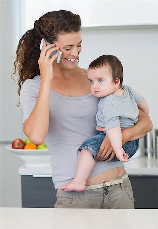 Smiling mother using cellphone while carrying baby boy in kitchen at home Stock Photo - Budget Royalty-Free & Subscription, Code: 400-07335659