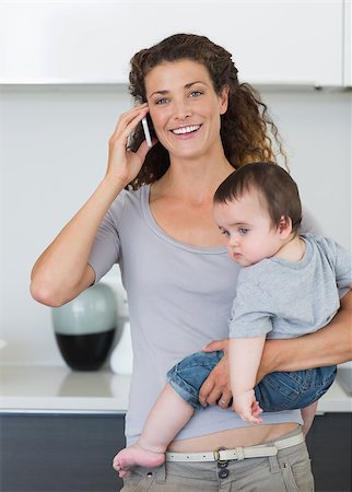 Happy woman answering smartphone while carrying baby boy in kitchen Stock Photo - Budget Royalty-Free & Subscription, Code: 400-07335660