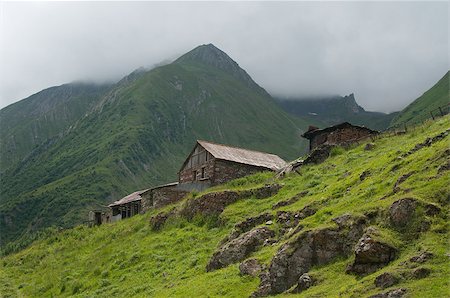 Old stone house is tucked among the mountains (Georgia).  Green bright grass and stones are in the foreground, mountains covered with clouds are in the background. Foto de stock - Super Valor sin royalties y Suscripción, Código: 400-07415246