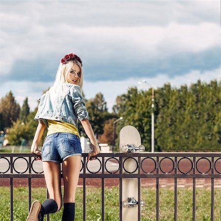 Blond girl in short shorts and denim coat with roses wreath on head turns back having climbed fence with skateboard hung on it Foto de stock - Super Valor sin royalties y Suscripción, Código: 400-07509935