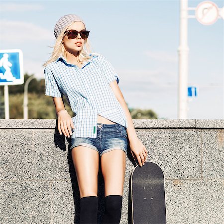 simsearch:400-07506274,k - Smiling blond girl in short shorts, checked sleeveless shirt, black stockings and sunglasses leans on granite fence next to skateboard Photographie de stock - Aubaine LD & Abonnement, Code: 400-07509952