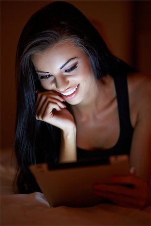 simsearch:400-05364625,k - Young woman reading a tablet in the darkness smiling as the glow from the screen illuminates her beautiful face Foto de stock - Super Valor sin royalties y Suscripción, Código: 400-07771021